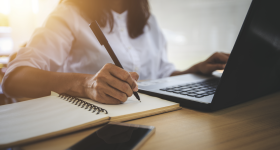 A woman writing at a desk alongside computer